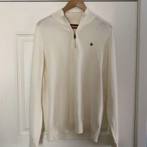 merinoulltröja från Morris - Säljer en stilren vit/kräm färgad tröja från Morris i 100% merinoull. Tröjan har en dragkedja vid halsen och ett broderat emblem på bröstet. Perfekt för en elegant och bekväm look.