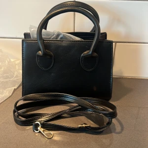 Svart handväska med axelrem - Snygg svart handväska i skinn med avtagbar axelrem. Perfekt för att bära det viktigaste med stil. Väskan har två handtag och en dragkedja upptill för säker förvaring.