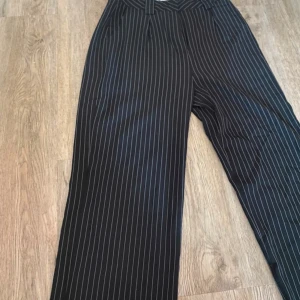 Svarta randiga byxor från NAKD - Snygga svarta byxor med vita ränder från NAKD. Byxorna har en klassisk design med hög midja och vida ben, vilket ger en stilren look. Perfekta för en chic och elegant stil.