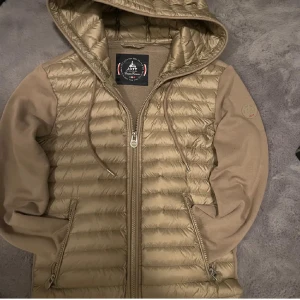 Beige quiltad cardigan med huva från JOTT - Säljer en beige quiltad jacka från Jott med huva och dragkedja. Jackan har långa ärmar och en stilren design. Perfekt för vår och höst väder. 