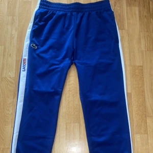 Lacoste byxor - Blå Lacoste byxor helt ny och oanvända, beställde fel storlek och dem har bara varit i garderoben. Byxorna har en elastisk midja och är perfekta för sportiga aktiviteter. Lacoste-loggan är tryckt i rött och blått längs sidan.