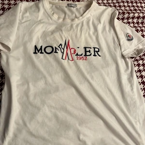 Vit t-shirt från Moncler - Säljer en stilren vit t-shirt från Moncler med broderad logga i svart och rött på bröstet. T-shirten har korta ärmar och en rund halsringning. Perfekt för en avslappnad look.