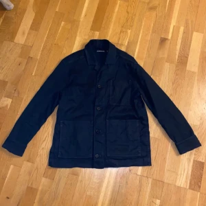 Blå owershirt från J.Lindeberg - Snygg blå owershirt från J.Lindeberg i storlek S. Jackan har en klassisk design med knappar framtill och stora fickor. Perfekt för en stilren look.