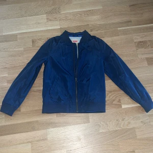 Blå bomberjacka från Levi's - Snygg blå bomberjacka från Levi's med broderad logga på ryggen. Jackan har en dragkedja framtill och ribbade muddar vid ärmslut och nederkant. Perfekt för en cool och avslappnad stil.