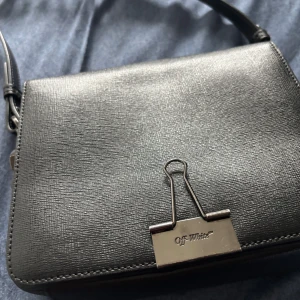 Svart axelväska från Off-White - Snygg svart axelväska från Off-White med en unik klämma som stängning. Väskan har en stilren design med justerbar axelrem och en praktisk innerficka med dragkedja. Perfekt för att bära dina nödvändigheter med stil.