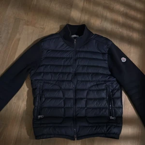 Svart pufferjacka från Moncler - Snygg svart pufferjacka från Moncler med dragkedja och ribbad krage. Jackan har långa ärmar och ett stilrent märke på ärmen. Är storlek M men känns mer som S 