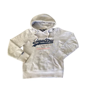 Grå hoodie från SuperDry - Säljer en stilren grå hoodie från SuperDry med tryck framtill. justerbar huva med snören. Perfekt för en avslappnad look. Storlek: XS. HÖR AV ER VID FRÅGOR😊