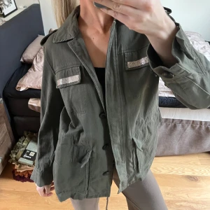 Olivgrön jacka från Zara - Snygg olivgrön jacka från Zara med långa ärmar och dekorativa detaljer på bröstfickorna. Jackan har en klassisk krage och flera praktiska fickor framtill. Perfekt för en stilren look.