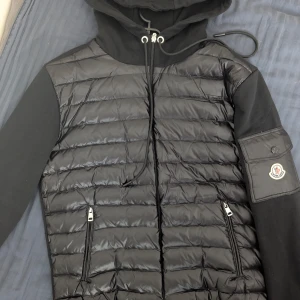 Originell Moncler Cardigan (Ny) - Med NFC såklart! Snygg svart jacka från Moncler med quiltad design och huva. Jackan har dragkedja framtill och två praktiska fickor med dragkedjor. Moncler-logotypen finns på ärmen.  !!! 90% Goose Down !!!    PRIS SÄNKT 3.5k till 2.4k