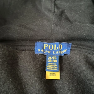 Svart hoodie från Ralph Lauren - Säljer en svart hoodie från Ralph Lauren med dragkedja och huva. Den har en liten röd logga på bröstet och praktiska fickor framtill. Perfekt för en avslappnad stil. Använd max 4 gånger och är helt stilren. 