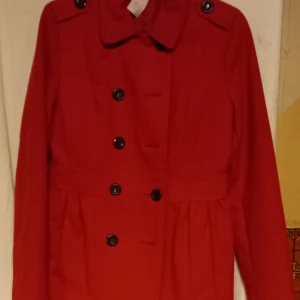 Röd fodrad finkappa från Divided - Snygg röd fodrad finkappa från Divided med knappar framtill och en klassisk krage. Perfekt för att ge en färgklick till din outfit. Kappan har långa ärmar och en stilren design som passar till många tillfällen.