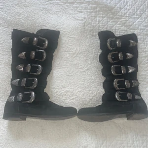 Svarta boots med spännen - Snygga svarta boots i skinn med flera dekorativa spännen i silver. Stövlarna har en dragkedja på insidan för enkel på- och avtagning. Tillverkade i Italien, dessa boots är perfekta för en stilren look. Nypris 8000kr.