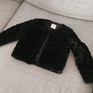 Svart pälsjacka från Lindex Kids - Säljer en supermjuk och stilren svart pälsjacka från Lindex Kids. Perfekt för att hålla sig varm och snygg under kyliga dagar. Jackan har långa ärmar och en enkel design som passar till alla outfits. Perfekt för höst och vinter!