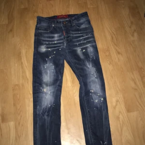 Mörkblå jeans med slitningar - Säljer ett par mörkblå jeans från DSQUARED2 med coola slitningar och färgstänk. De har en klassisk femficksdesign och en knappgylf. Perfekta för en avslappnad och trendig look. Passar både till vardags och fest!