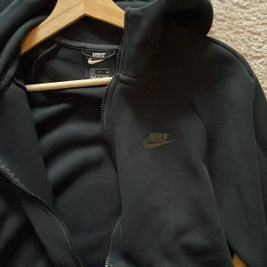Svart Nike tech hoodie - Säljer en svart hoodie från Nike i  bra skick. First Season drop från 2019. Klassiska Nike tech. Den har en dragkedja framtill och en praktisk huva. Perfekt för att sätta ihop en dress eller använda som singel. Passar både till träning och vardagsbruk. Storlek S