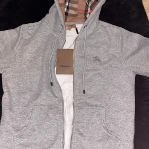 Burberry kofta - Säljer en stilren grå huvtröja från Burberry med klassiskt rutigt mönster i huvan. Tröjan har dragkedja framtill och en broderad logotyp på bröstet. Perfekt för en avslappnad look. Helt oanvänd. L men passar också M