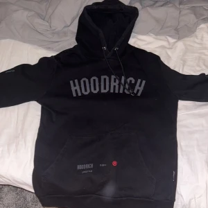 Svart hoodie från Hoodrich - Säljer en svart hoodie från Hoodrich med tryck på bröstet och ärmen. Den har en klassisk känguruficka och justerbar huva. Perfekt för en avslappnad stil. Som ny inga tecken på användning äkta