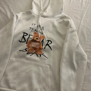 Vit hoodie med nalledekor - Superfin vit hoodie med en söt nalledekor på framsidan. Texten 'IT'S A TEDDY BEAR' ger den en unik touch. Perfekt för mysiga dagar eller som en statement piece i garderoben. Den har en klassisk känguruficka och en bekväm huva med dragsko. Ganska tunn hoodie så inte så himla varm, passar bäst på sommaren när man är lite kylig💕