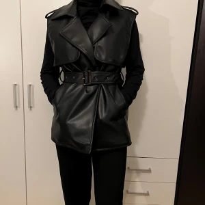 Svart skinnväst med bälte - Snygg svart skinnväst med bälte i midjan. Perfekt för en cool och stilren look. Västen har en klassisk krage och är ärmlös, vilket gör den idealisk för lager-på-lager-styling. Passar både till vardags och fest.