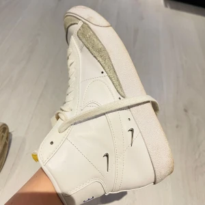 Vita Nike sneakers - Säljer ett par vita Nike sneakers i skinn med snygg snörning och en högre modell. De har en stilren design med små silverdetaljer och en robust sula. Perfekta för både vardag och festliga tillfällen. Skorna är i mycket bra skick och redo för nya äventyr! 