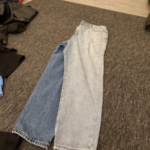 Tvåfärgade jeans - Säljer ett par unika tvåfärgade jeans med en ben i ljusblått och det andra i mörkblått. De har en klassisk femficksdesign och är tillverkade i denim. Perfekta för dig som vill sticka ut med en cool stil!