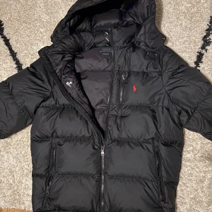 Svart dunjacka från Polo Ralph Lauren - En praktiskt sätt ny Polo jacka, inga slitningar, inga hål och inga defekter. Jackan är i storlek L och tillkommer med en justerbar midja, fungerar utmärkt för storlek M om man spänner åt midjan. Nypris på denna jacka ligger runt 4500-4899 kr, medans jag säljer för 3499.                          Skriv gärna vid intresse