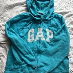 Turkos hoodie från GAP - Säljer en snygg turkos hoodie från GAP med dragkedja och stor logga på framsidan. Perfekt för en avslappnad stil. Den har en mysig huva och långa ärmar.