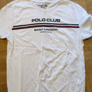 Vit t-shirt från Polo Club Saint Vincent - Säljer en vit t-shirt från Polo Club Saint Vincent med tryck i svart och rött. T-shirten har en rund halsringning och korta ärmar. Perfekt för en avslappnad stil.
