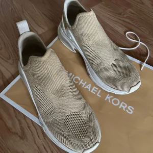 Beige sneakers från Michael Kors - Säljer ett par snygga beige sneakers från Michael Kors i stickat material. De har en modern slip-on design utan snörning, vilket gör dem superenkla att ta på och av. Perfekta för en avslappnad och stilren look. Använda men i bra skick.