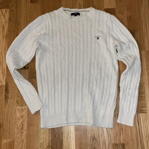 Vit stickad tröja från Gant - Säljer en stilren vit stickad tröja från Gant. Den har ett klassiskt kabelstickat mönster och en liten broderad logga på bröstet. Perfekt för både höst och vinter, och den är supermjuk och bekväm. Passar till alla tillfällen!