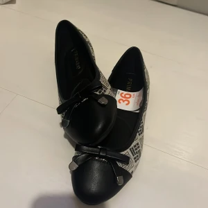 Svart och vit ballerinasko från Primark - Snygga ballerinaskor i storlek 36. De har ett svart och vitt mönster med en elegant rosett på tån. Perfekta för både vardag och festliga tillfällen. Skorna är i mycket bra skick då de inte använts en enda gång och har prislappen kvar. Skorna passar storlek även storlek 38 då de är stora i storleken. 