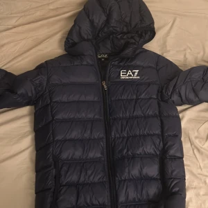Mörkblå dunjacka från Emporio Armani - Säljer en mörkblå dunjacka från Emporio Armani EA7. Jackan har en dragkedja och är quiltad för extra värme. Den har tre små hål, 2 st längst ner vid armen och 1 på axeln. Det går att fixa med lite silver tejp eller sy ihop hålen. Kontakta mig vid minska lilla fundering!