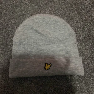 Grå mössa från Lyle & Scott  - Säljer en snygg grå mössa från Lyle & Scott. Den är stickad och har en broderad gul logga framtill. Perfekt för att hålla sig varm under kyliga dagar. Passar både till vardags och lite mer uppklädda tillfällen. (Accepterar endast Swish pga lurendrejerier) (Checka gärna in andra kläder på min profil för att köpa tillsammans för ett rabatterat pris) 