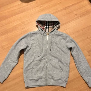 Grå hoodie från Burberry - Säljer en stilren grå hoodie från Burberry med klassiskt rutigt mönster i huvan. Tröjan har dragkedja och snörning vid huvan. Perfekt för en avslappnad och trendig look.