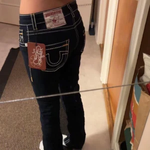 Mörkt blåa jeans från True Religion W30 - Snygga svarta jeans från True Religion med kontrasterande vita sömmar och klassiska bakfickor. Jeansen har en tight passform och låg midja, perfekt för en stilren look. Märkeslappen syns tydligt på baksidan.