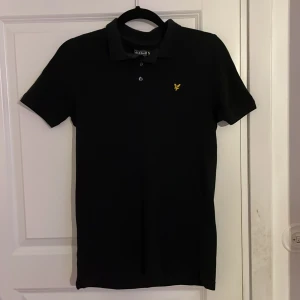 Svart pikétröja från Lyle & Scott - Snygg svart pikétröja från Lyle & Scott med klassisk krage och knappar. Den har en gul broderad logotyp på bröstet. Perfekt för en stilren look. 14/15 years