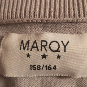 Grå tröja från MarQy - Säljer en stilren beige tröja från MarQy i storlek 158/164. Tröjan har en v-ringad hals och är broderad med MarQy-loggan på bröstet. Perfekt för både vardag och festliga tillfällen. Den är i mycket bra skick och redo för en ny ägare! 
