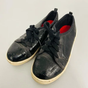 Svarta sneakers från H&M - Snygga svarta sneakers från H&M i storlek EUR 29. De har en glansig yta med snörning och en vit sula. Insidan är röd vilket ger en cool kontrast. Perfekta för vardagsbruk och ger en stilren look. Använda men i bra skick.