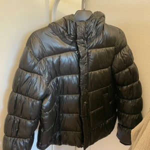 Svart pufferjacka från Superdry - Säljer en svart pufferjacka från Superdry i storlek EU 40. Jackan skulle jag säga är lite liten i storlek. Vanligtvis hade jag haft 38 och den sitter perfekt fast den är i 40. Jackan är i  bra skick och perfekt för vintern. Den har en huva och stängs med dragkedja. Superdry-loggan finns på ärmen. Grym för kalla dagar! Jackan har ett litet hål på ena armen som är limmat.