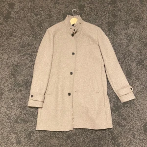 Beige kappa med knappar - Elegant beige kappa med hög krage och knappar framtill. Den har långa ärmar med knappdetaljer vid ärmsluten och en stilren design. Perfekt för kyligare dagar. Köpt för 1400 men enbart testad. 