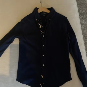 Mörkblå skjorta från Polo Ralph Lauren - Säljer en snygg mörkblå skjorta i mjukbomull från Polo Ralph Lauren. Skjortan har långa ärmar och knappar framtill. Den är i ett klassiskt snitt och passar perfekt för både vardag och fest. Materialet känns mjukt och skönt mot huden. Perfekt för höst och vår!