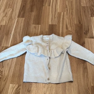 Beige kofta med volang från H&M - Söt beige kofta från H&M med volangdetaljer längs axlarna. Perfekt för att hålla de små varma under kyligare dagar. Koftan har knappar framtill och är tillverkad i ett mjukt material.