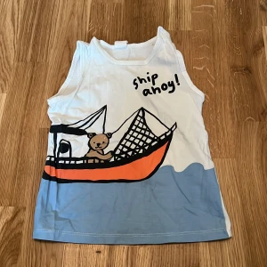 Vit ärmlös topp med tryck från H&M - Säljer en söt vit ärmlös topp från H&M med ett tryck av en nalle i en båt och texten 'ship ahoy!'. Toppen har blå och orange detaljer och är perfekt för varma dagar.