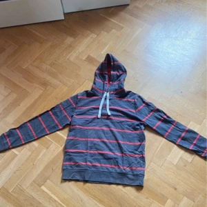 Grå och röd randig hoodie från Gant - Säljer en snygg grå hoodie från Gant med röda ränder och justerbar huva. Perfekt för en avslappnad stil. Den har långa ärmar och en klassisk passform.