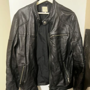 Svart Premium skinnjacka från Jack & Jones - Säljer en snygg svart skinnjacka från Jack & Jones i storlek S. Jackan har en cool bikerstil med dragkedjor på fickorna och en ståkrage med knapp. Perfekt för höst och vår, och den ger en snygg look till vilken outfit som helst.