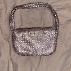 Glittrig axelväska från Bershka - Glittrig axelväska från Bershka med dragkedja. Väskan har en glittrande yta och en rosa insida.