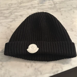 Svart ribbad mössa från Moncler - Snygg svart ribbad mössa från Moncler med deras ikoniska logotyp framtill. Perfekt för att hålla värmen under kyliga dagar.