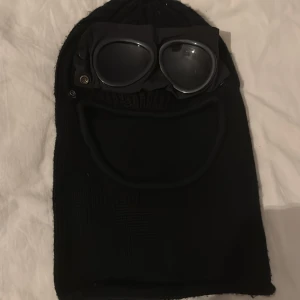 Svart stickad mössa med glasögon - Cool svart stickad mössa från C.P. Company med inbyggda glasögon. Perfekt för att hålla sig varm och se stilren ut. Mössan har en unik design med glasögon som kan fällas ner över ansiktet.