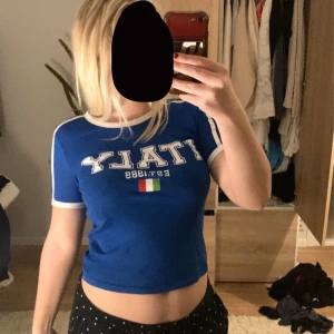 Blå croppad t-shirt med text - Säljer en blå ny croppad t-shirt med texten 'ITALY Est. 1899' och en liten italiensk flagga på framsidan. T-shirten har vita detaljer vid ärmslut och hals. Perfekt för en avslappnad sommarlook eller för att visa lite kärlek till Italien! 💙