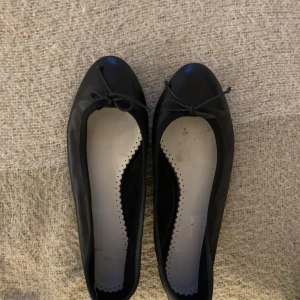 Svarta ballerinaskor med rosett - Eleganta svarta ballerinaskor i syntetmaterial med en söt rosett på tån. Perfekta för en stilren och bekväm look.